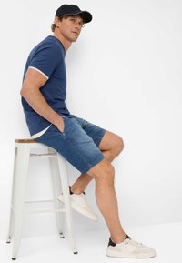 T-shirt blu navy a maniche corte con bordature bianche, abbinato a pantaloni corti di jeans. Scarpe da ginnastica chiare e un cappellino blu navy completano l'outfit.