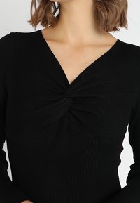 Top lungo a maniche nere a coste con scollatura a V e dettaglio annodato al petto per contrasto di texture e design.