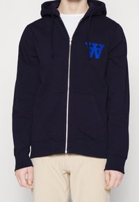 Navy zip-up hoodie made of cotton blend, features a kangaroo pocket, drawstring hood, and a blue embroidered logo on the chest.