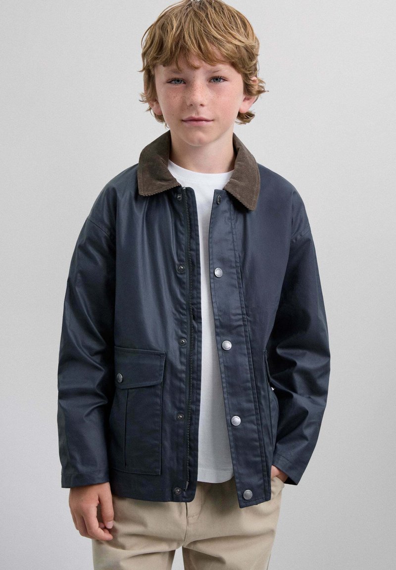 Niño con cabello castaño claro que lleva una chaqueta azul marino con cuello marrón, camisa blanca y pantalones beige, de pie contra un fondo gris claro.