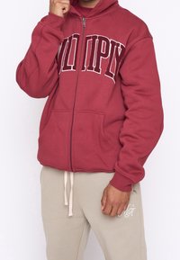 Sudadera con cremallera color marrón burdeos con texto bordado, que presenta un bolsillo frontal y puños acanalados. Llevada con pantalones de chándal de un ligero color beige.