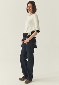 Femme souriante, debout avec les mains dans les poches, portant un haut blanc à manches longues, un jean foncé, un pull rayé noué autour de la taille et des chaussures marron.