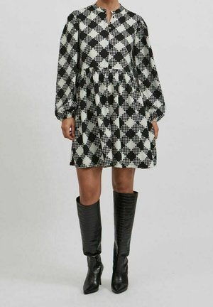 Kobieta w czarno-białej sukience w kratkę z długim rękawem i marszczoną talią, w połączeniu z czarnymi, teksturowanymi kozakami za kolano.