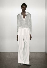 Lichtgrijze, lange mouwen shirt met een kraag, gedragen over een witte, hoog-waisted wijde broek, aangevuld met zilveren instapschoenen.