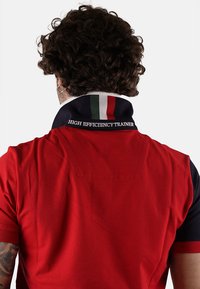 Polo rosso con accenti blu navy e colletto rigato in verde, bianco e rosso. "ALLENATORE AD ALTA EFFICIENZA" ricamato sul colletto.