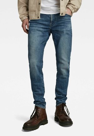 Jeans de mezclilla azules con un corte slim, que presentan áreas descoloridas y detalles de costura clásica. Combinados con botas de cuero marrón hasta el tobillo.