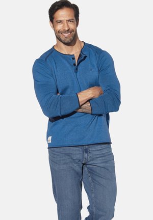 Blue long-sleeve Henley shirt with three buttons, contrasting black collar, and side tag, paired with light blue jeans.