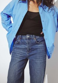 Šviesiai mėlyna mygtukais užsegama marškinėliai sluoksniuoti ant juodos maikės, derinami su aukštu juosmeniu, tamsiai mėlynais džinsais su matomu siūliniu.