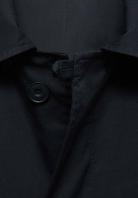 Cappotto nero con chiusura a bottone. Il tessuto appare liscio con una leggera lucentezza. Presenta un colletto a punta e dettagli di cucitura puliti.