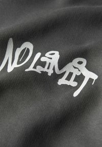 Sivá tkanina s hladkou textúrou, na ktorej je biely text v graffiti štýle, ktorý hovorí "ŽIADNE LIMITY." Dizajn je výrazný a abstraktný.