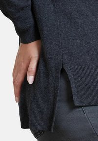 Mörkgrå tröja gjord av mjukt, strukturerat stickat tyg. Har ribbade ärmslut och sidokilar vid fållen. Hand som vilar försiktigt på tyget.