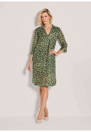 Blonde Frau steht, trägt ein grünes knielanges Kleid mit schwarzem und braunem Leopardenmuster und beigefarbene spitze High Heels, eine Hand in der Hüfte.