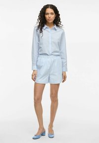 Chemise rayée bleu clair avec col à revers et fermeture à boutons, associée à un short bleu clair assorti. Des chaussures à talons bleues complètent la tenue.