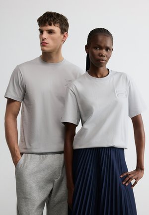 Junger Mann und junge Frau tragen hellgraue T-Shirts; Mann in Jogginghose und Frau in einem marineblauen Faltenrock, stehend vor einfarbigem Hintergrund.