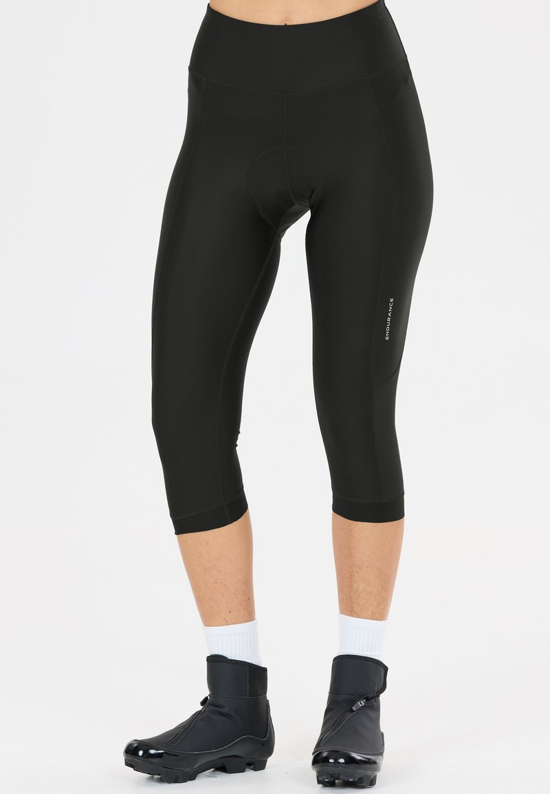 Schwarze, gepolsterte Rad-Caprihose, getragen mit weißen Socken und schwarzen Radschuhüberzügen, gezeigt an einer stehenden Person.