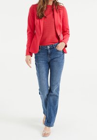 Röd figursydd kavaj över en texturerad röd topp, tillsammans med blå raka jeans; modell står med ena handen i fickan, iförd vita sandaler.