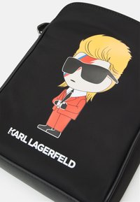 Černá taška s karikaturou postavy v červeném obleku, černých rukavicích, brýlích a blond vlasy, s červeným a modrým pruhem na tváři, text "Karl Lagerfeld" pod tím.