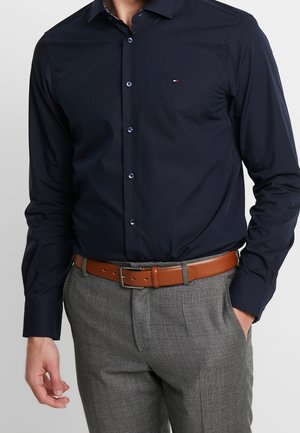 Mand iført en figursyet mørkeblå skjorte med knapper, stoppet ned i grå dressbukser med et lysebrunt læderbælte, den ene hånd i lommen.