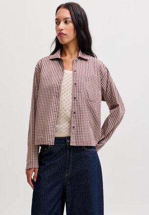 Femme portant une chemise à carreaux rouge et beige boutonnée par-dessus un haut en maille crème et un jean large foncé, regardant sur le côté avec une main sur la hanche.