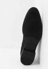 Zapato de ante negro con suela de goma plana. La suela presenta un patrón de agarre texturizado y un talón ligeramente elevado. Tamaño marcado como 42.