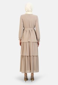 Beige volangklänning med långa ärmar och midja med bälte. Tillverkad av lättviktsmaterial, med en liten knappstängning i ryggen.