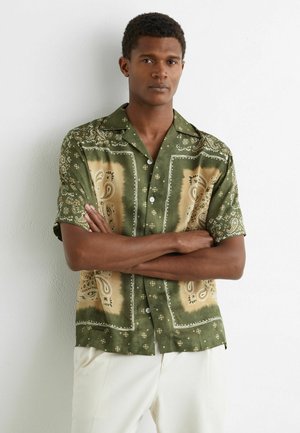 Mand med korslagte arme iført en kortærmet skjorte med grønne og beige mønstre og hvide bukser, stående op ad en hvid væg.