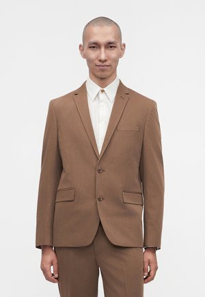 Uomo che indossa una giacca da completo marrone e pantaloni abbinati con una camicia bianca abbottonata, in piedi davanti a uno sfondo bianco uniforme.
