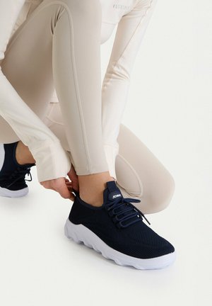 Person in beige activewear kneeling and adjusting navy blue sneaker with white sole on a white background.