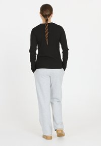 Schwarzes langärmliges Rippoberteil, graue Jogginghose und beiges Slipper. Haare zu einem Zopf gestylt. Weißer Hintergrund. Rückansicht.