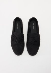Svarta mocka slip-on skor med en knutdetalj på ovansidan, texturerad yta och en platt gummisula. Logotyp inuti skon.