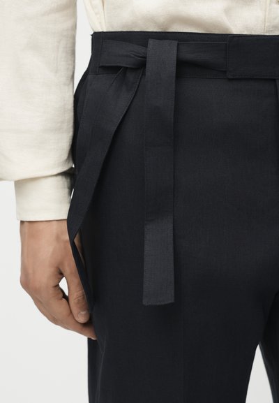 Χέρι που φοράει μπεζ μακρυμάνικο πουκάμισο δίπλα σε μαύρο ψηλόμεσο παντελόνι με δεμένη υφασμάτινη ζώνη.