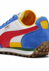 Adidașii Puma Easy Rider cu partea superioară din material sintetic alb, accente din piele de căprioară albastră, dungi roșii, guler galben și talpă texturată maro.
