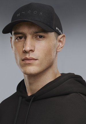 Man with serious expression wearing a black hoodie and black cap with "HYROX" logo on front and side against a gray background.