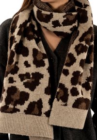 Écharpe beige avec un motif léopard en marron foncé et marron clair. Texture tricotée avec un ourlet côtelé. Portée par-dessus un cardigan gris foncé.