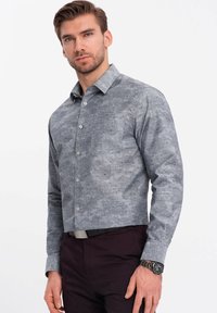 Camisa gris de botonadura con un patrón floral, que presenta un cuello estructurado, mangas largas y un corte ajustado. Se lleva con pantalones oscuros.