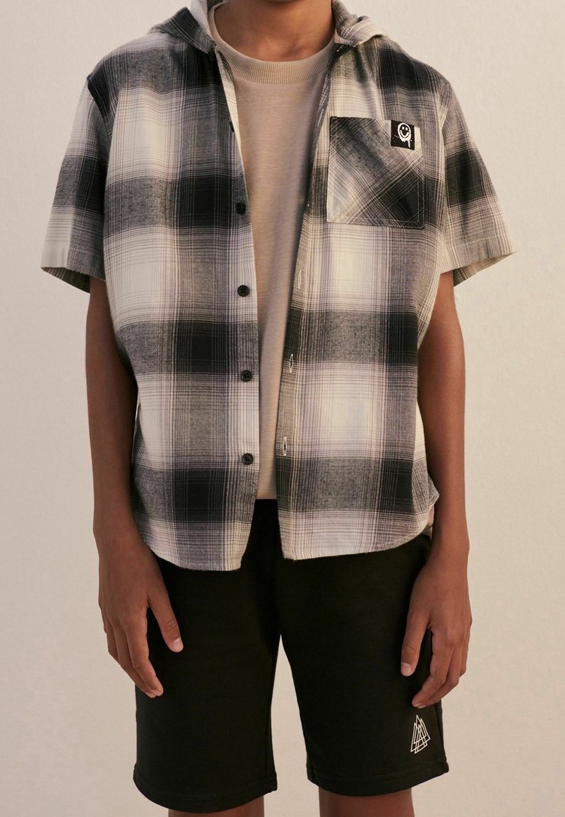 Personne portant une chemise à manches courtes à carreaux noirs et blancs boutonnée par-dessus un t-shirt beige, associée à un short noir avec un petit logo blanc.