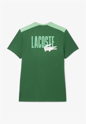 Zaļš kokvilnas T-krekls ar gaiši zaļiem plecu akcentiem. Aizmugurē ir liels "LACOSTE" teksts un krokodila grafika. Īsas piedurknes.