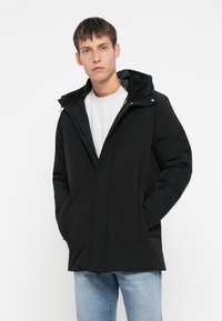 Svart pufferjakke med hette, med en glatt ytterside og en krage foret med fleece. Glidelås foran og sidelommer.