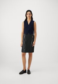 Colete sem mangas em azul marinho com um tecido texturizado, apresentando um bolso frontal e botões, combinado com uma saia lápis em pele sintética preta com detalhes em zíper.