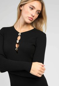 Haut en tricot noir côtelé avec un col rond, comportant trois boutons décoratifs le long d'une ouverture en clé verticale. Manches longues.