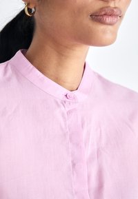 Femme portant une chemise rose clair à col mandarin avec le bouton supérieur fermé et une boucle d'oreille anneau doré à l'oreille gauche.
