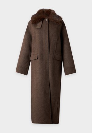 Femme aux cheveux bouclés portant un long manteau marron foncé avec un col en fourrure et des mocassins noirs, debout devant un fond clair uni.