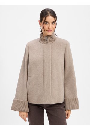 Frau trägt eine beige Wolljacke mit geripptem hohem Kragen und breiten Manschetten, kombiniert mit dunkelbraunen Hosen vor weißem Hintergrund.