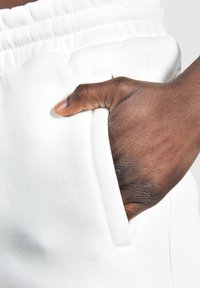 Hvide sweatpants med elastisk talje og sidelommer, fremhæver et tekstureret stof og en hånd indsat i lommen.