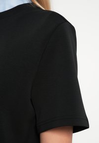 Κοντινό πλάνο ενός ατόμου που φοράει ένα απλό μαύρο κοντομάνικο μπλουζάκι, δείχνοντας τον ώμο και τον άνω βραχίονα σε ανοιχτό φόντο.