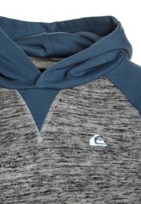 Sudadera de punto gris con acentos azules en el cuello y los hombros. Presenta un diseño triangular en la parte frontal y un pequeño logo en el pecho.