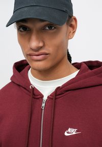Sweatshirt fechado cor bordô feito de algodão, com um logotipo branco da Nike e cordões. A pessoa que o está usando também tem um boné preto.