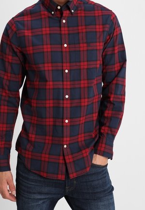 Mann trägt ein rot-navyblau kariertes Hemd mit langen Ärmeln und dunkle blaue Jeans, eine Hand in der Tasche.