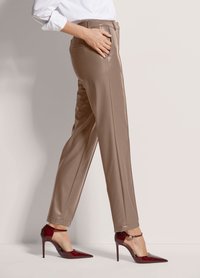 Vrouw in taupe hoge leren broek en bordeauxrode lakleren hoge hakken, zijwaarts staand met één hand in de zak.