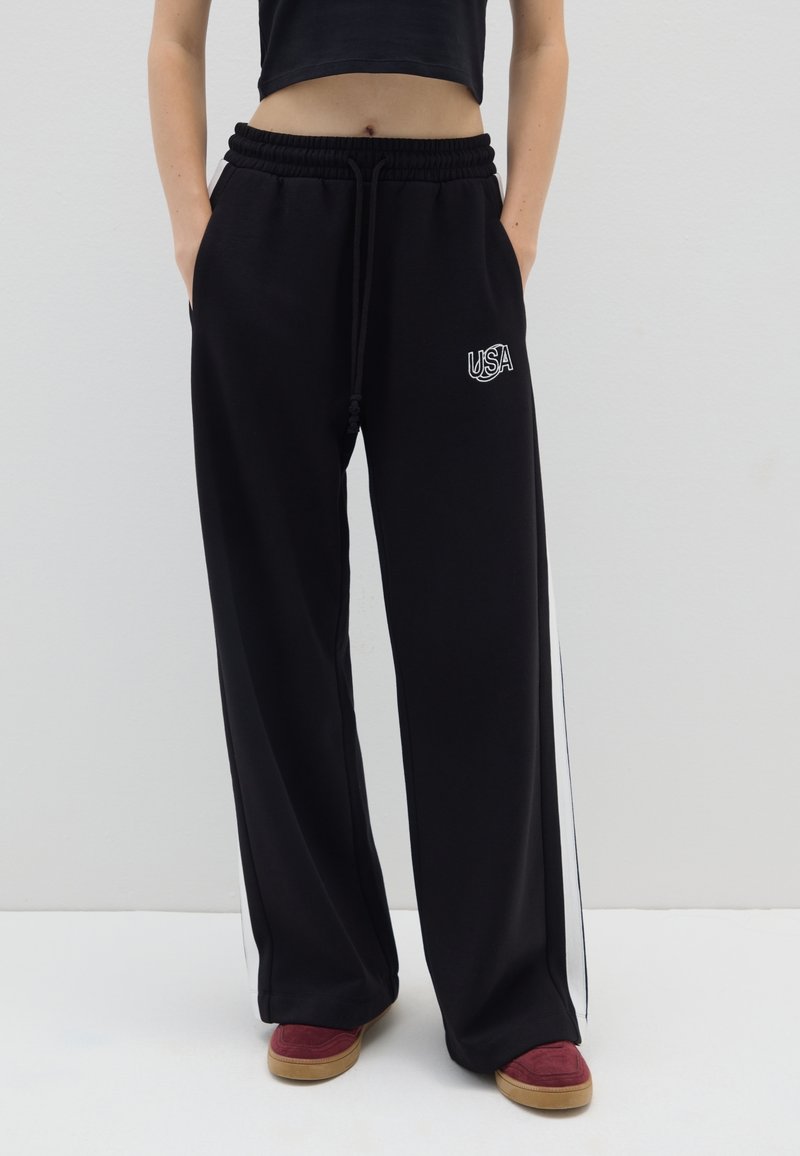 Pantalones de chándal negros con un corte holgado, cintura elástica, rayas blancas a los lados y un logo bordado de "USA" en la parte delantera derecha.